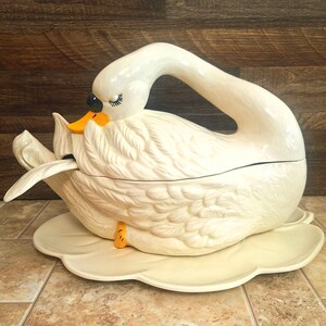 May include: A cream-colored ceramic swan-shaped tureen with a matching lid and base. The swan features a long, curved neck handle, a bright orange beak, and painted black eyes with eyelashes. A white ceramic ladle rests inside the tureen.