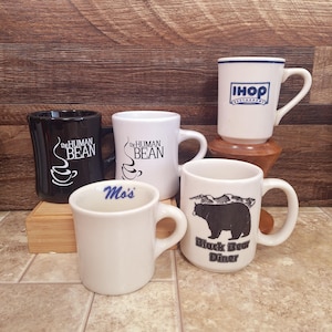 May include: A collection of five ceramic mugs featuring various logos and designs. Included are a black and a white mug with The Human Bean logo, a white IHOP Restaurant mug, a white mug with the text Mo's, and a white mug with a black bear graphic.