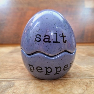 May include: A purple ceramic salt and pepper shaker set shaped like an egg. The top half of the egg is labeled "salt" and the bottom half is labeled "pepper".