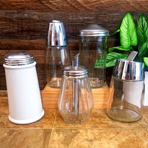 May include: A collection of vintage glass and metal sugar dispensers and shakers. The dispensers have clear glass bodies with various shapes and silver-colored metal lids. One shaker is white ceramic with a silver lid. The background is a wooden surface.