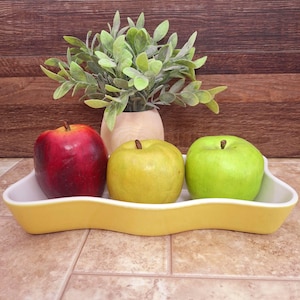 May include: A yellow ceramic serving dish with a wavy design holds three apples: a red, a yellow, and a green one. A small vase with green leafy plant sits behind the apples. The background is a brown wooden wall.