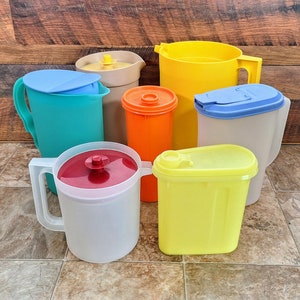 May include: A collection of six colorful plastic pitchers in various sizes and shapes. The pitchers are in shades of blue, green, yellow, orange, and white.