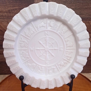 May include: White glass plate with a ruffled edge and embossed zodiac symbols around a compass rose design. The plate is displayed on a black metal stand, showcasing its intricate details and vintage aesthetic. A decorative piece for display.