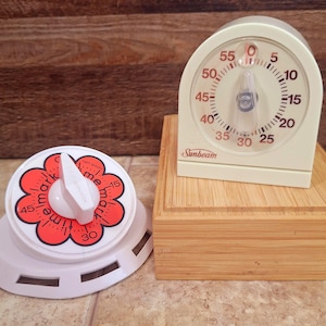May include: Two vintage kitchen timers. One is a white, flower-shaped timer with a red floral design and the words "mark me time". The other is a cream-colored timer with a dial and the brand name "Sunbeam".