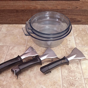 May include: A clear glass baking dish with a blue rim and handles, stacked with another similar dish. Four metal and black pan lifters are arranged in front of the dishes. The lifters have a triangular shape.