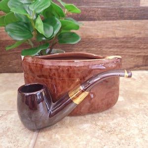 May include: A brown ceramic tobacco pipe with a dark brown bowl and stem, accented with gold-colored bands. The pipe rests in front of a brown ceramic ashtray and a small potted plant with green leaves.