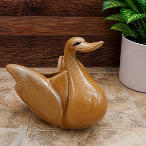 May include: A glossy, light brown ceramic planter shaped like a swan. The swan has a smooth, reflective finish with a curved neck and a hollowed-out body designed to hold items. The piece features a stylized, elongated beak and wings.