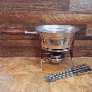 May include: A vintage fondue set with a stainless steel pot, wooden handle, and stand. The set includes a burner and several fondue forks. The pot is approximately 8 inches in diameter.