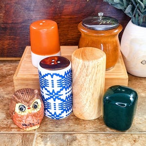 May include: A collection of five salt and pepper shakers. The shakers are made of different materials, including wood, ceramic, and glass. The shakers are arranged on a wooden surface. One shaker is a brown ceramic shaker with a white band and three brown dots. Another shaker is a white ceramic shaker with a blue and white geometric pattern. A third shaker is a brown wooden shaker with a silver top. A fourth shaker is a light brown wooden shaker. The fifth shaker is a dark green ceramic shaker.