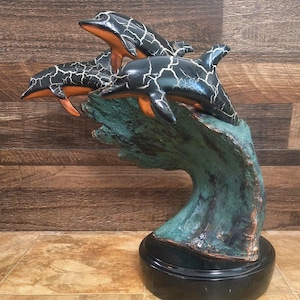 May include: A sculpture of three dolphins leaping from a wave. The dolphins are black with white crackle patterns and orange accents. The wave is teal and the base is black, creating a striking contrast.