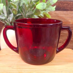 May include: A ruby red glass mug with two handles. The mug has a rounded shape and a smooth, glossy finish. The handles are curved and attached to the sides of the mug. The mug is sitting on a wooden surface.