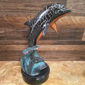 May include: A ceramic dolphin sculpture with a black and white crackle finish. The dolphin has orange accents and is mounted on a teal and black base. The sculpture is on a black circular base.