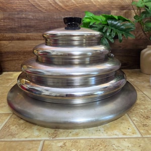 May include: A stack of five circular stainless steel pot lids of varying sizes, arranged from largest at the bottom to smallest at the top. Each lid has a smooth, reflective metallic finish and a rounded edge. A small black knob sits on the top lid.