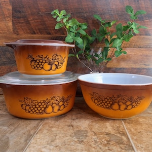 May include: Three brown and white ceramic mixing bowls with a grape and fruit design. The bowls are nested together. The largest bowl is a mixing bowl, the middle bowl is a casserole dish with a lid, and the smallest bowl is a small serving bowl.