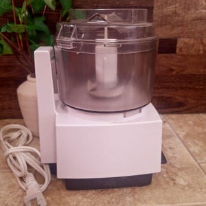 May include: A white electric food processor with a clear plastic bowl and lid. The appliance features a central white blade assembly inside the bowl, a white base, and a white power cord with a standard plug extending from the side of the unit.
