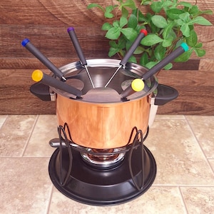 May include: A copper fondue pot with a black base and stand. The pot has a silver lid with slots for fondue forks. The forks have black handles and colored tips: yellow, blue, purple, red, and green. The pot is on a tiled surface.