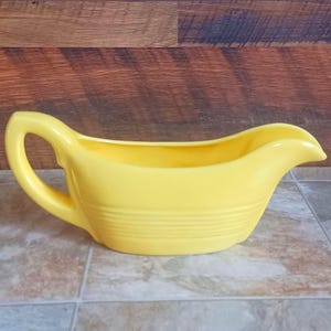 May include: A bright yellow ceramic gravy boat with a handle and a spout. The boat has horizontal ridges around the base and is set against a neutral background. This vintage-style serving dish is ideal for sauces and gravies.
