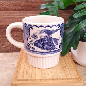 May include: A cream-colored ceramic mug featuring a blue transferware-style illustration of a steam locomotive train. The mug has a scalloped border design around the upper rim and a vertical ribbed texture on the lower half of the exterior body.