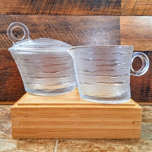 Peut inclure: Un ensemble de deux pots à crème et à sucre en verre transparent avec un motif texturé ondulé. Les pots sont posés sur une base en bois.