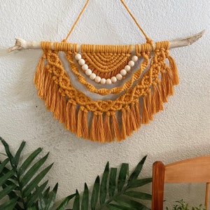 May include: A mustard yellow macrame wall hanging suspended from a light-colored wooden branch. The design features intricate knotted patterns, a row of smooth round wooden beads, and long fringe tassels hanging in a semi-circular arrangement.