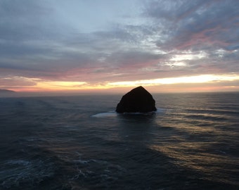 May include: A large, dark rock formation rises from the ocean, silhouetted against a colourful sunset sky. The sky is a mix of pink, orange, and purple hues, with clouds reflecting the light. The water is a deep blue, with waves gently rolling.