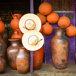 May include: A collection of terracotta pots and vases in various sizes and shapes, some with a dark brown glaze. The pots are arranged on a wooden surface with a purple wall in the background. There are also two straw hats in the image.