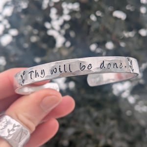 May include: A silver cuff bracelet with the phrase "Thy will be done." stamped on the front. The bracelet has a hammered texture and small leaf designs on each end of the text. A silver ring with a floral design is also visible.