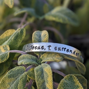 May include: A silver bracelet with the inscription "O Jesus, I surrender all" on a green leafy background.