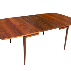 May include: A brown wooden dining table with a rectangular top and four slender legs. The table has a leaf that extends the length of the table.