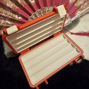 Peut inclure: Un boîtier de rangement rectangulaire ouvert avec un extérieur orange vif et un intérieur blanc comportant quatre canaux horizontaux moulés. Le boîtier comprend deux fermetures à loquet blanches sur le bord supérieur et une charnière. Le texte STYLE A est visible.