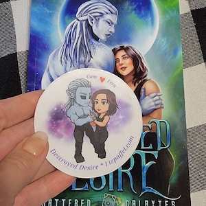 May include: A book cover with the title "Destroyed Desire" by Liz Paffel, featuring a fantasy illustration of two characters. A round sticker with cartoon characters is in front of the book. The background is a black and white checkered pattern.