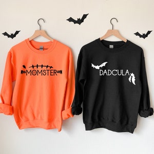 May include: Two orange and black Halloween themed sweatshirts. The orange sweatshirt has the text "MOMSTER" with a stitched line above it. The black sweatshirt has the text "DADCULA" with two bats above it.