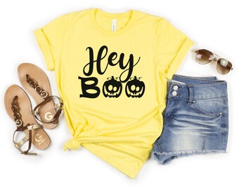 May include: Yellow short-sleeved t-shirt with the text "Hey Boo" in black script with two black pumpkin faces. The shirt is paired with denim shorts and sandals.