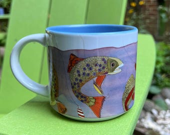 May include: A ceramic mug with a light blue interior and a white exterior featuring a colorful illustration of a trout fish with orange fins and blue spots. The design includes a small red and white striped fishing lure hanging near the fish.