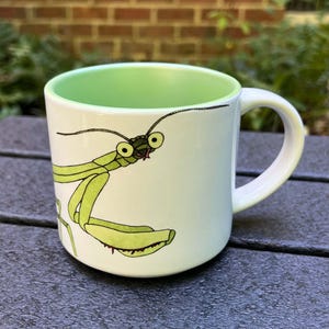 May include: A white ceramic mug with a green rim and a hand-painted illustration of a praying mantis. The mantis is green with black eyes and is facing the viewer.