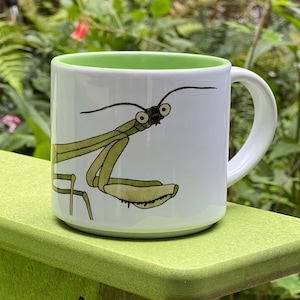 May include: A white ceramic mug featuring a cartoon illustration of a green praying mantis with large, wide-set eyes. The mug has a solid light green interior and a matching handle, providing a clean contrast to the white exterior surface.