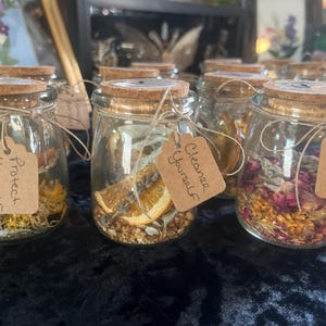 May include: Glass jars with cork lids, filled with dried herbs, flowers, and spices. Each jar has a tag with phrases like "Protect Yourself", "Cleanse Yourself", and "Love Yourself". Tied with twine.