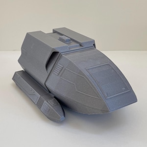 May include: A gray, 3D-printed model of a futuristic spacecraft. The model features a rectangular body with angled sides and a small, raised section on top. Two smaller, cylindrical components extend from the sides. The overall design suggests a science fiction theme.