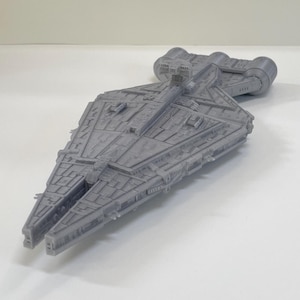 May include: A gray, detailed model of a starship with a long, angular body and multiple protrusions. The model has intricate surface details, including panels and ridges, and is set against a plain white background. The ship's design suggests a science fiction or fantasy theme.