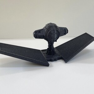 May include: A black 3D printed model of a TIE fighter with a detailed design. The model has two wings that are folded down.