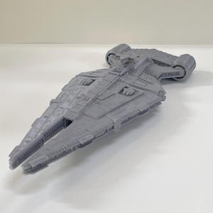 Op de afbeelding: Een grijs, gedetailleerd model van een ruimteschip met een lange, hoekige romp en ingewikkelde oppervlakte details. Het model heeft twee cilindrische motoren aan de achterkant en een gedetailleerde cockpit. Het ruimteschip is een verzamelobject.