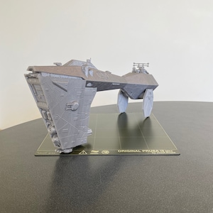 Op de afbeelding: Een grijs 3D-geprint model van een ruimteschip, met een gedetailleerd ontwerp en ingewikkelde kenmerken. Het model staat op een zwart, getextureerd oppervlak met de tekst "ORIGINAL PRUSA i3 MK3 by Josef Prusa".