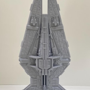 Op de afbeelding: Grijs, 3D-geprint model van een ruimteschip. Het model heeft een gedetailleerd, getextureerd oppervlak met ingewikkelde geometrische ontwerpen. Het ruimteschip heeft een hoog, verticaal ontwerp met een brede basis en twee prominente verticale vinnen.