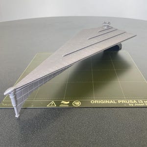 Puede incluir: Un modelo gris impreso en 3D de una nave espacial Star Destroyer sobre una superficie verde texturizada. La superficie tiene una señal de advertencia y el texto "ORIGINAL PRUSA i3 MK3S by Josef Prusa".