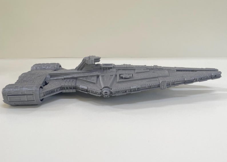 Op de afbeelding: Een grijs, gedetailleerd model van een ruimteschip, met ingewikkelde ontwerpen en een getextureerd oppervlak. Het ruimtevaartuig heeft een lange, slanke romp met diverse uitsteeksels en een duidelijk voorste gedeelte. Het model is waarschijnlijk een verzamelobject.