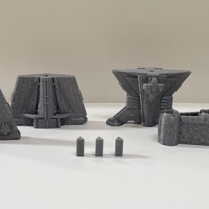 Op de afbeelding: Een verzameling grijze, geometrische 3D-geprinte structuren. De set bevat twee driehoekige stukken, een veelzijdig gebouw, een tafelachtige structuur, een kasteelachtige muur en drie kleine obeliskvormige stukken. De objecten staan op een wit oppervlak.