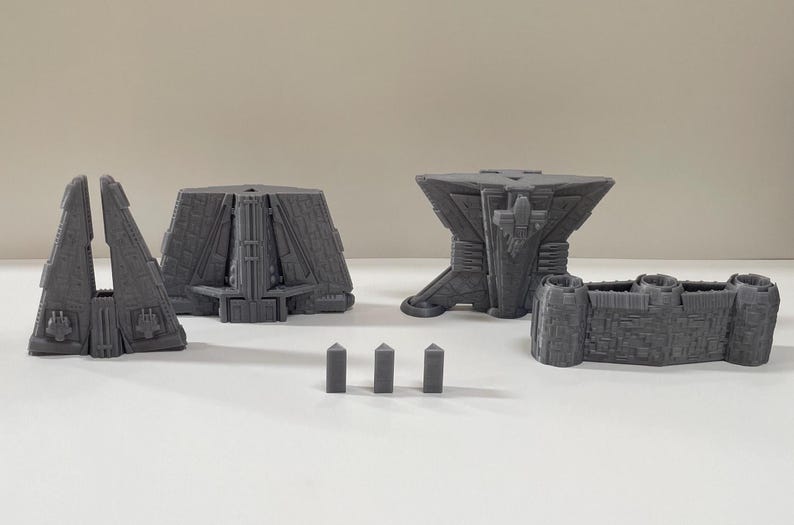 Op de afbeelding: Een verzameling grijze, geometrische 3D-geprinte structuren. De set bevat twee driehoekige stukken, een veelzijdig gebouw, een tafelachtige structuur, een kasteelachtige muur en drie kleine obeliskvormige stukken. De objecten staan op een wit oppervlak.