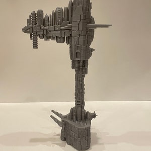 May include: A grey 3D printed model of a large, detailed spaceship with a long, vertical tower and multiple levels. The spaceship has a rounded top and a base with three legs.