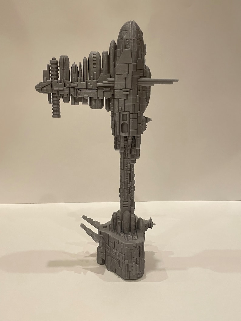 May include: A grey 3D printed model of a large, detailed spaceship with a long, vertical tower and multiple levels. The spaceship has a rounded top and a base with three legs.