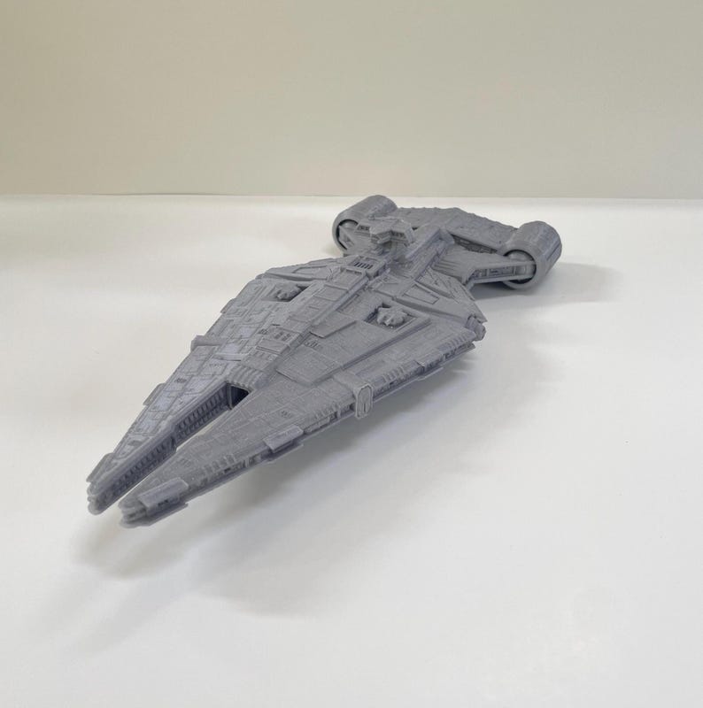 Op de afbeelding: Een grijs, gedetailleerd model van een ruimteschip met een lange, hoekige romp en ingewikkelde oppervlakte details. Het model heeft twee cilindrische motoren aan de achterkant en een gedetailleerde cockpit. Het ruimteschip is een verzamelobject.